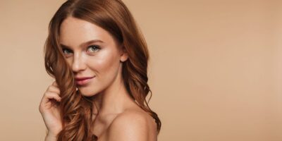 Beauty portrait of mystery smiling ginger woman with long hair posing sideways and looking at the camera over cream background